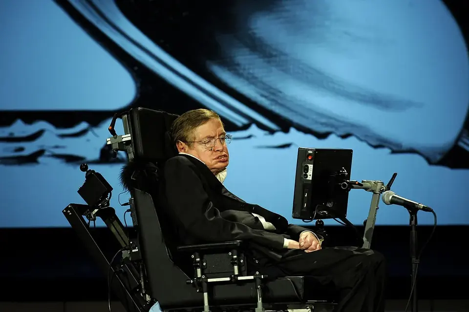 Stephen Hawking dans son fauteuil sur une scène, face à un grand écran, lors d'une conférence publique