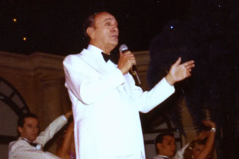 Tino Rossi en costume blanc chantant au micro sur une scène, bras tendu, entouré de danseurs